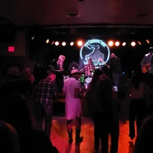 People dancing and watching a live band performing on a dimly lit stage with a circular band logo in the background